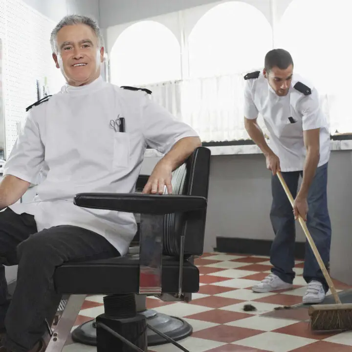 10018582 Portrait Of Happy Senior Male Barber With Apprentice Sweeping Floor At Barbershop 720x720 